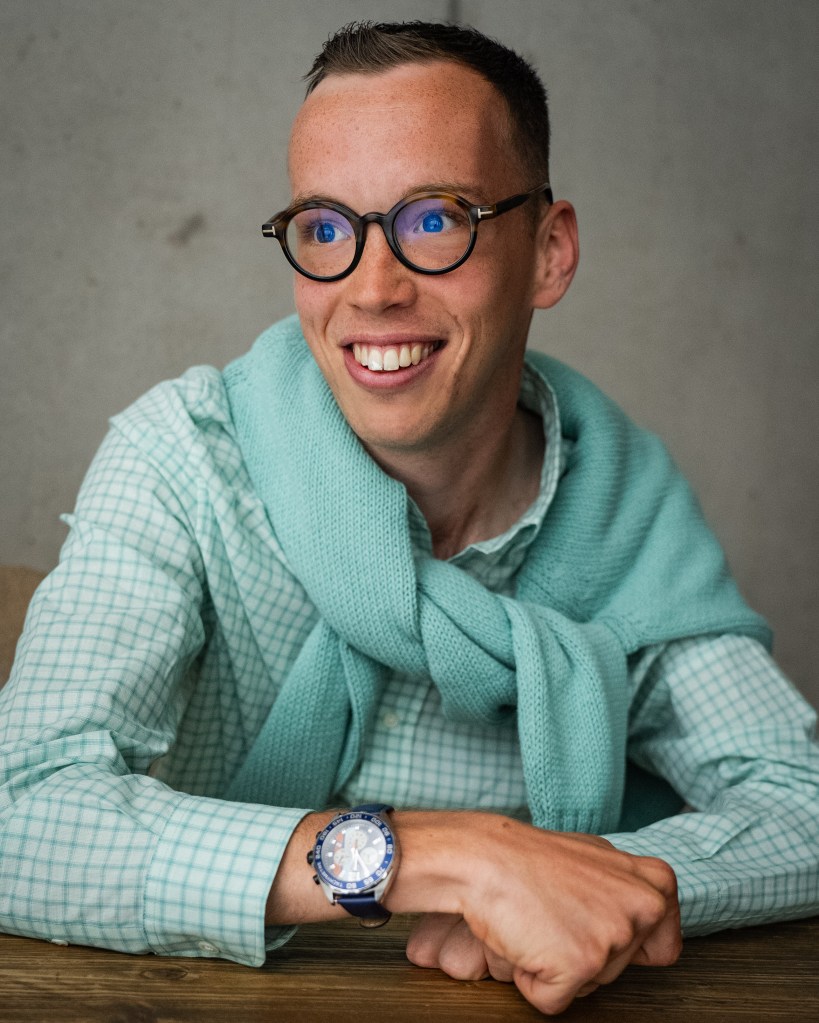 Keron, a white man with gelled brown hair, is sat with his arms learning onto a brown, wooden table. He is looking to his right, smiling. He is wearing a turquoise, checked shirt with a turquoise woollen jumper wrapped around his neck. He is also wearing brown glasses and a blue watch.